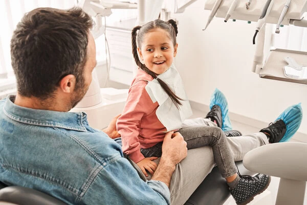 Young girl smiling in dental chair with father during family dentistry visit