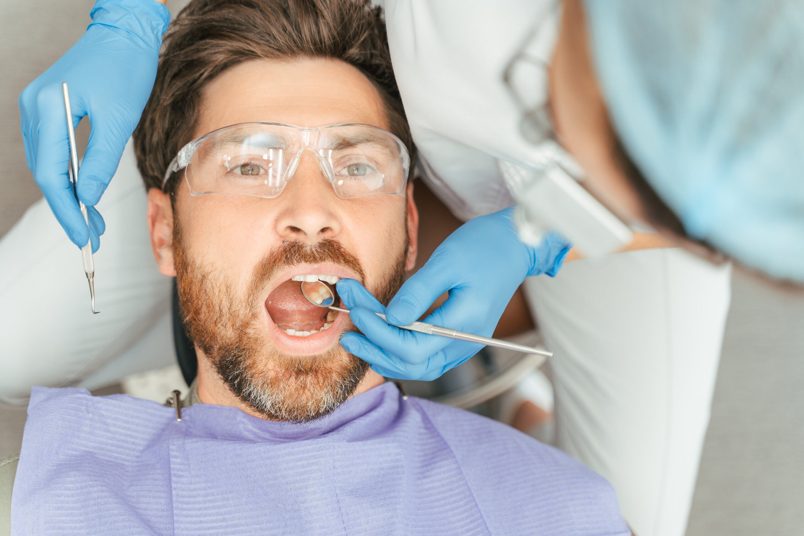 Portrait of handsome bearded man patient sitting in dental chair wearing protective glasses with open mouth, closeup. Female dentist treats teeth with instruments. Concept of dental care